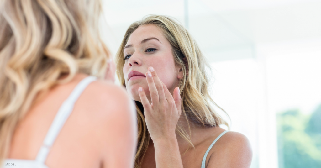 Woman examining her skin in the mirror after a treatment of dermal fillers.