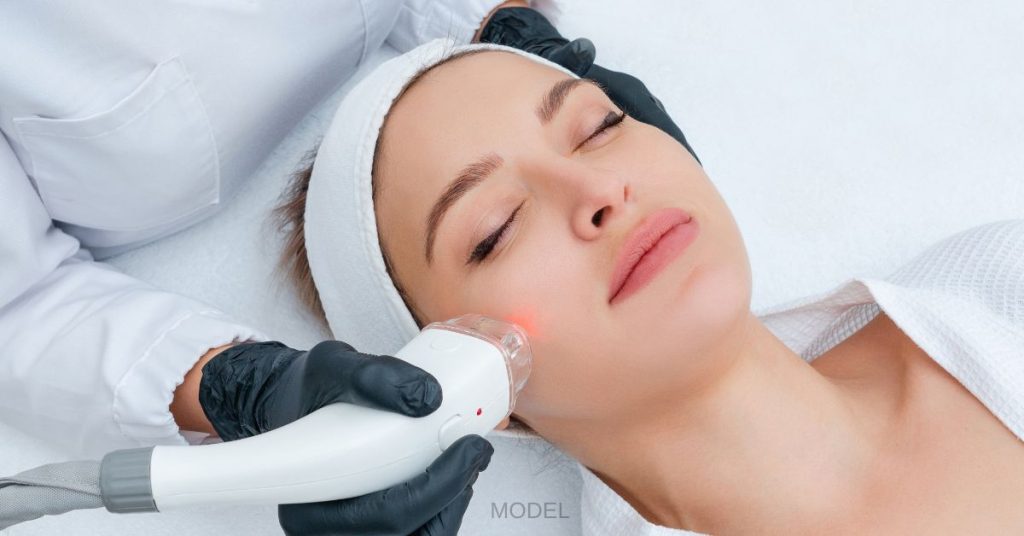 White woman (model) with her eyes closed, wearing a white headband, and receiving a laser skin treatment by someone wearing black gloves.
